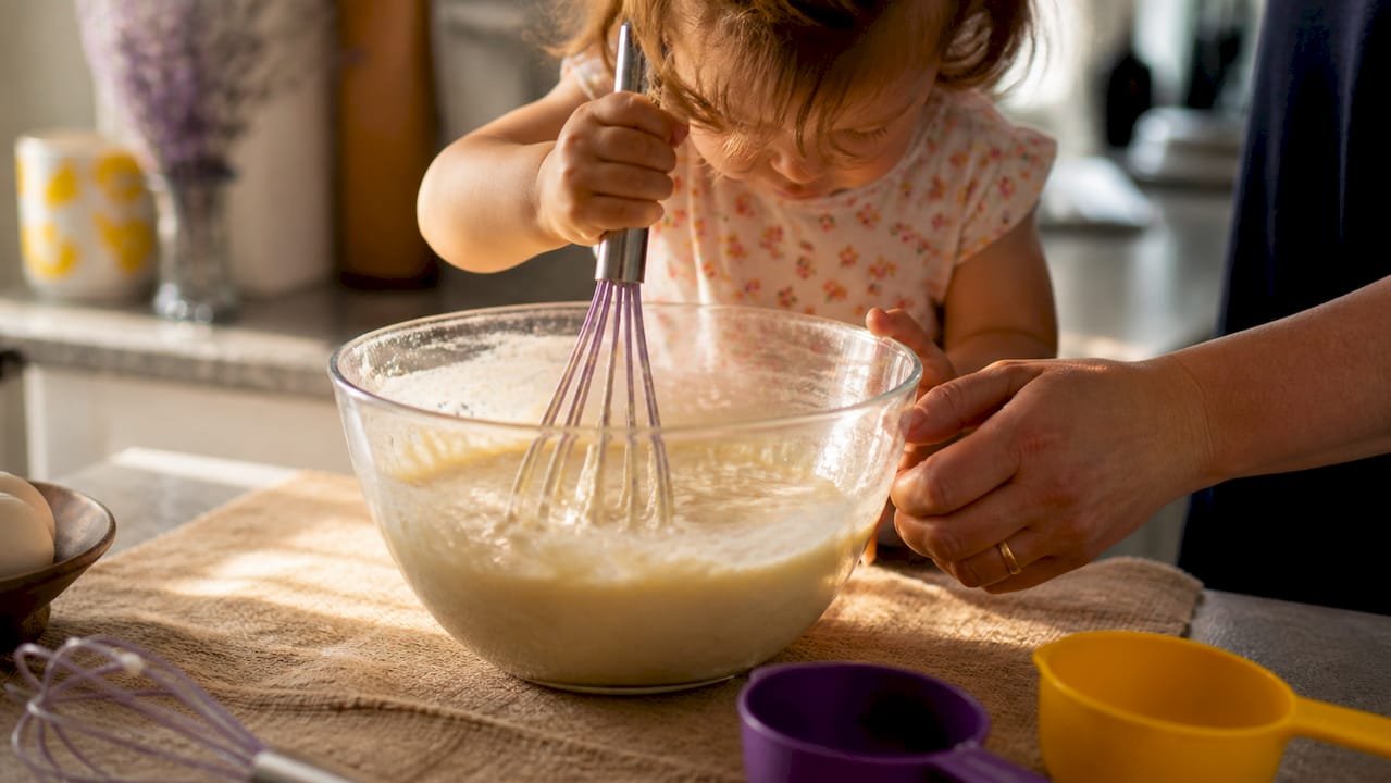 Toddler stirring pancake batter with adult hand support